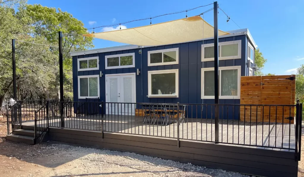 Elevated wooden deck and patio with black metal railing, outdoor furniture, shade sail, and a blue tiny home.
